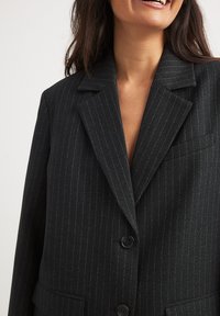 Blazer noir à rayures avec un design structuré, doté d'un col cranté, de deux boutons et d'une poche externe. Détail de tissu texturé visible.