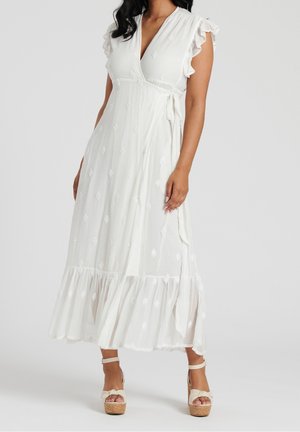 Femme portant une robe portefeuille blanche longue jusqu'aux chevilles avec des manches à volants et des sandales compensées, debout sur un fond uni.