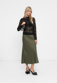 Woman wearing a black lace top, olive green midi skirt, black bow detail heels, and carrying a small black handbag.