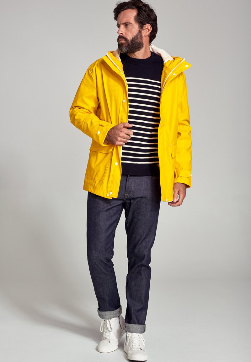 Gul vandtæt jakke med hætte, flad front og tryklåse, lagt over en marineblå og hvidstribet sweater og mørke jeans. Hvide sneakers.