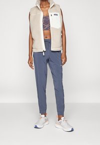 Beige fleece vest with a high collar, white zip pocket; patterned sports bra underneath, gray tapered pants, and light sneakers.