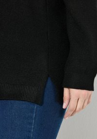 Hand ruht an der Seite, trägt einen schwarzen Strickpullover und blaue Jeans vor einem schlichten Hintergrund.