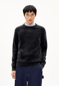 Schwarzer Rundhals-Pullover mit strukturiertem Finish, ausgestattet mit Raglanärmeln und gerippten Bündchen. Über einem hellblauen Hemd mit Knopfleiste getragen.