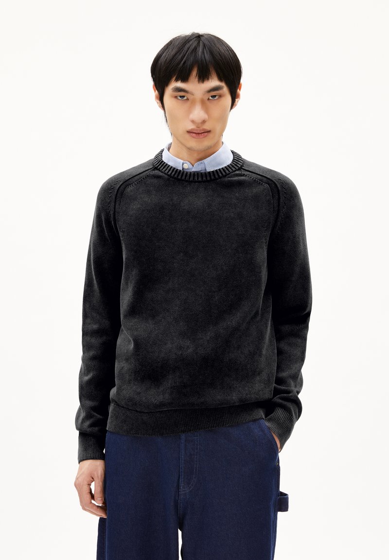 Schwarzer Rundhals-Pullover mit strukturiertem Finish, ausgestattet mit Raglanärmeln und gerippten Bündchen. Über einem hellblauen Hemd mit Knopfleiste getragen.