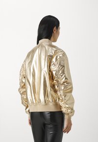Metallisch goldene Bomberjacke mit gerafften Seitendetails, geripptem Saum und Bündchen. Der Stoff hat eine glatte, glänzende Textur.