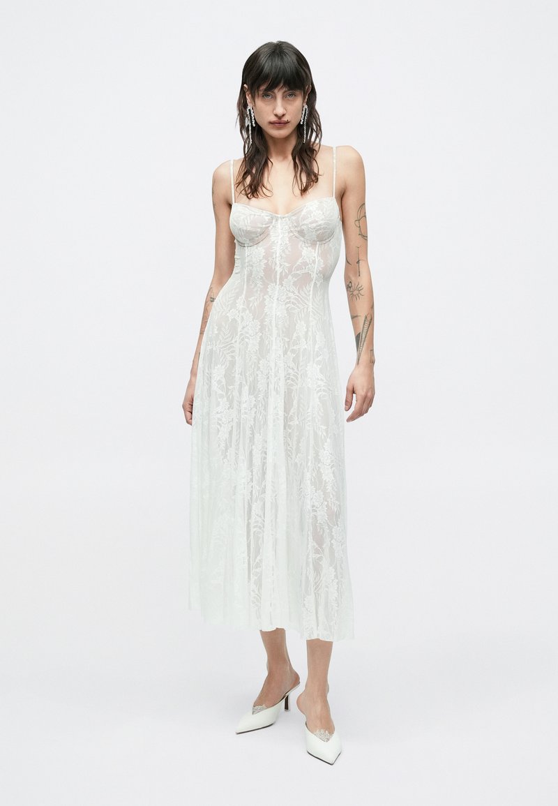 Femme aux cheveux foncés portant une robe midi blanche en dentelle transparente avec des fines bretelles et des talons blancs pointus, debout devant un fond uni.