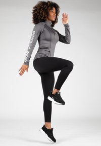 Grijze sportjack met rits en logo op de mouw, gecombineerd met zwarte leggings en zwarte sneakers, in een actieve houding poserend tegen een neutrale achtergrond.