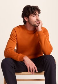 Man with dark curly hair wearing an orange sweatshirt and black pants, sitting on a wooden chair, resting his chin on his hand, looking right.