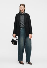 Γυναίκα που φορεί μαύρο σακάκι με διπλό πέτο, γκρι πουλόβερ με ψηλό γιακά, μπλε τζιν παντελόνι με φαρδιά μπατζάκια, καφέ ψηλοτάκουνα, κρατώντας μαύρη τσάντα σε απλό φόντο.