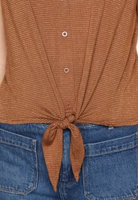 Bruine top met korte mouwen, verticale strepen en een strik aan de voorkant bij de taille. Kenmerkt zich door knopen en een zachte textuur. Gecombineerd met spijkerbroek.