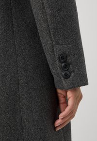 Mâneca unui palton gri din lână cu un finisaj texturat, având trei nasturi negri și o croială ajustată pentru un design structurat.