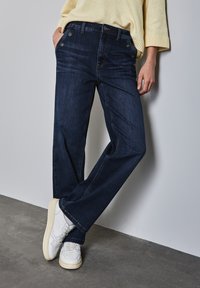 Jeans de mezclilla azul oscuro con corte recto, que cuentan con cinco bolsillos y herrajes de botón metálico. Combinados con zapatillas blancas.
