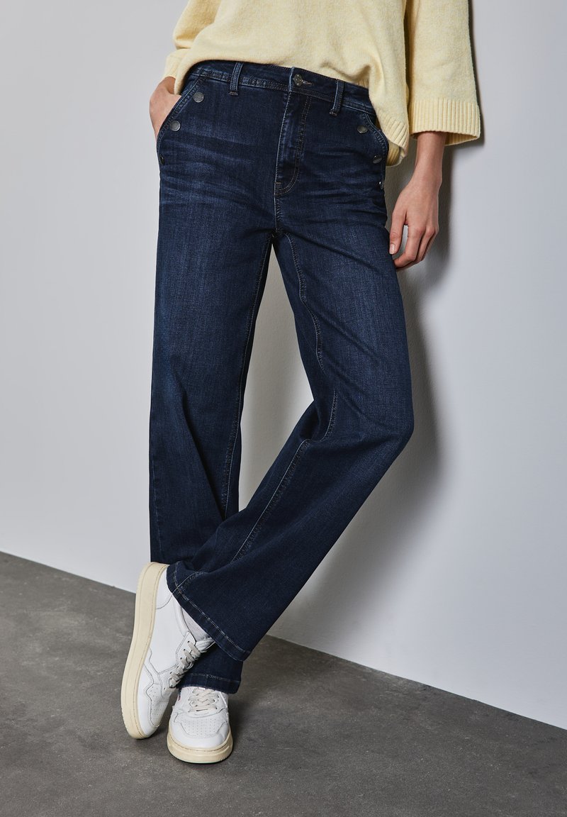 Jeans de mezclilla azul oscuro con corte recto, que cuentan con cinco bolsillos y herrajes de botón metálico. Combinados con zapatillas blancas.