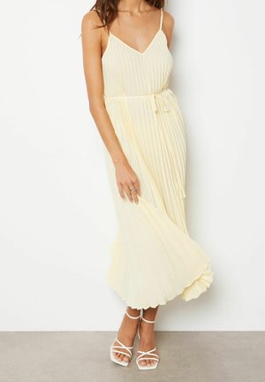 Femme portant une robe midi plissée sans manches, de couleur jaune pâle, avec de fines bretelles et des sandales blanches à talons hauts et lanières.