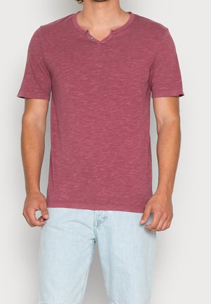 Homme portant un t-shirt henley à manches courtes couleur maroon avec deux boutons et un short en denim bleu clair, debout devant un fond uni.