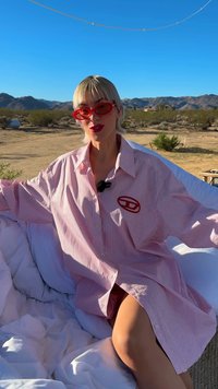 Person with short blonde hair wearing red sunglasses and an oversized red-striped shirt, sitting outdoors in a desert landscape.