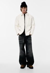 Valkoinen fleecetakki, jossa nappisuljenta ja taskut, tummat haalistuneet väljät farkut, ruskeat saappaat ja musta neulottu pipo.