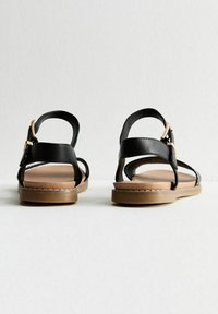Sandalias de cuero negro con correas ajustables en el tobillo, plantillas color tostado, suelas planas y un diseño minimalista con acentos de hardware en tono dorado.