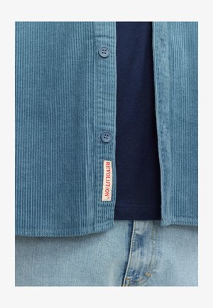 Lichtblauw corduroy shirt met ribbeltextuur, met twee blauwe knopen en een rode "REVOLUTION" label. Gecombineerd met een donkerblauwe T-shirt.