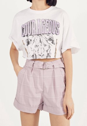 Kobieta nosząca biały krótki t-shirt z lwem i napisem "COURAGEOUS", w połączeniu z jasnofioletowymi szortami z wysokim stanem i paskiem.