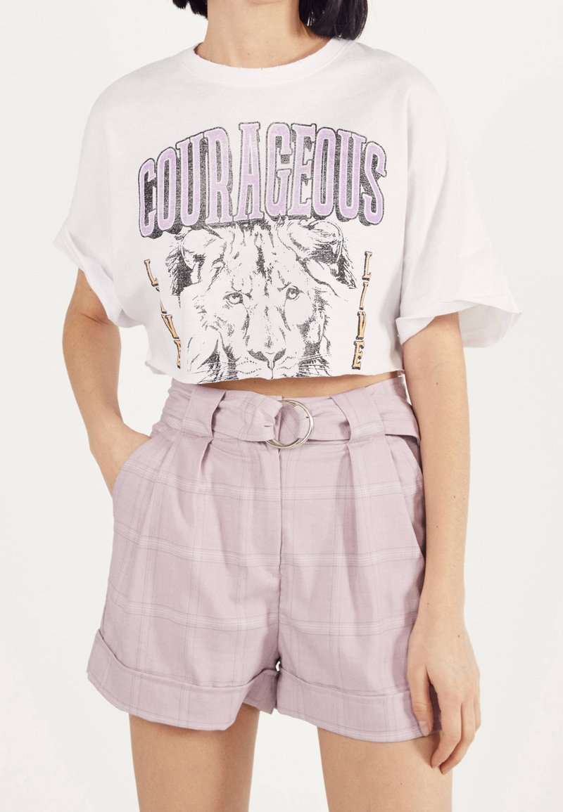 Femme portant un t-shirt court blanc avec un lion et le texte "COURAGEOUS", associé à un short taille haute violet clair avec ceinture.