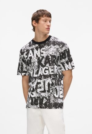 Młody mężczyzna w czarno-białym graficznym T-shircie z różnymi napisami, w połączeniu z białymi spodniami, stojący na białym tle.