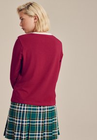 Punainen pitkähihainen paita pyöreällä pääntiellä, yhdistettynä vihreään ruudulliseen laskostettuun hameeseen, jossa on valkoisia ja keltaisia raitoja. Sileä pinta.