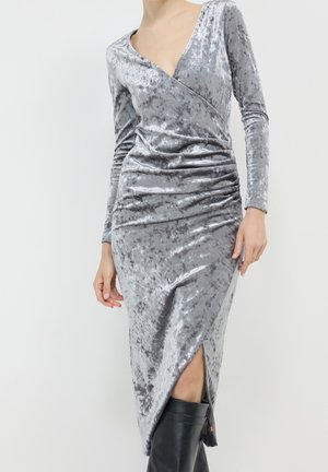 Frau in einem silbernen, zerknitterten Samt-Wickelkleid mit langen Ärmeln und einem Schlitz vorne, dazu schwarze kniehohe Stiefel, stehend vor weißem Hintergrund.