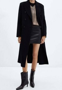 Nainen, jolla on päällään musta pitkä takki, beige puuvillaneuleliivi mustan paidan yllä, musta nahkainen mini-hame ja mustat korkeat nilkkasukat.