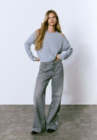 Camisa gris de manga larga con cortes en los hombros, combinada con unos pantalones vaqueros grises de pierna ancha con patrones de estrellas. Unos zapatos negros de punta completan el look.