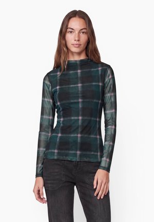 Femme aux longs cheveux bruns portant un haut à manches longues à carreaux vert foncé et rose et un pantalon noir, debout devant un fond blanc uni.
