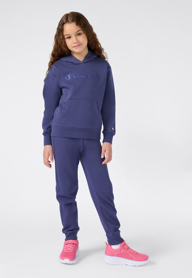 Ragazzina in piedi con felpa e pantaloni della tuta blu navy Champion, indossando sneakers fucsia su sfondo bianco.