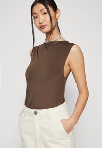 Brauner ärmelloser Bodysuit mit hohem Kragen und glatter Textur, kombiniert mit hellen Hosen, die einen Knopfverschluss und Taschen haben.