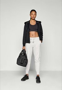 Mulher jovem em top desportivo preto, hoodie com fecho éclair preto, calças de treino brancas e sapatilhas pretas, segurando uma mala de ginásio preta, em pé contra um fundo simples.