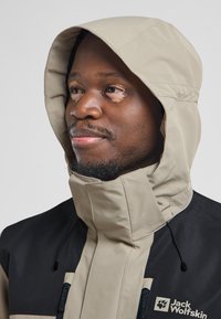 Giacca impermeabile beige e nera con colletto alto, cappuccio regolabile e tasche con zip. Logo visibile sul davanti.