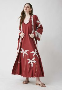 Robe maxi rouge avec broderie blanche en forme de palmier, dotée de bords fringés et de manches évasées. Le mannequin porte des sandales plates marron à enfiler.