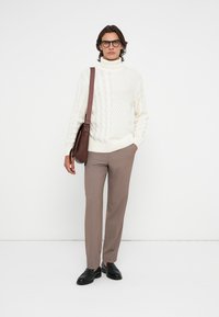 Weißer, kabelgestrickter Rollkragenpullover mit strukturierten Mustern, kombiniert mit braunen, maßgeschneiderten Hosen und schwarzen Slipschuhen. Braune Schultertasche.