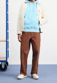 Persona con un hoodie ligero azul de Nike, una chaqueta de forro polar color crema, pantalones anchos marrones y zapatillas blancas, de pie al lado de un carrito de metal azul.