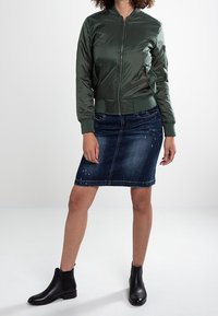 Zielona satynowa kurtka bomberka z zapięciem na zamek, ściągaczami przy rękawach, w połączeniu z ciemnoniebieską spódnicą ołówkową i czarnymi botkami za kostkę.