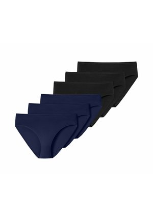 Donna con capelli ricci scuri che indossa un reggiseno nero e mutande nere a vita alta in piedi davanti a uno sfondo chiaro e uniforme.
