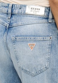 Τα ανοιχτό μπλε τζιν παντελόνια διαθέτουν ένα τρίγωνο σήμα "GUESS" στην πίσω τσέπη. Το ύφασμα έχει υφή με ξεθωριασμένη εμφάνιση και ραμμένες ραφές.