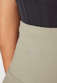 Dettaglio ravvicinato di una persona che indossa leggings beige Nike e una maglietta nera, con un focus sulla zona della vita dove è visibile il logo swoosh di Nike.