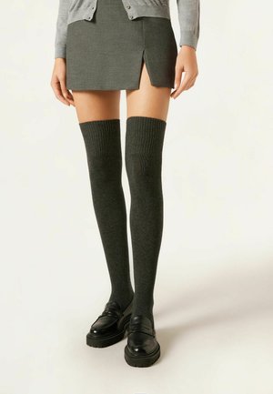 Chaussettes montantes grises foncées, tricotées avec des bords côtelés, associées à une jupe grise et des mocassins noirs, avec une légère fente sur le côté.