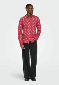 Man in een rode lange mouwen shirt met witte sterpatronen, zwarte wijde broek en zwarte schoenen, staand met één hand in zijn zak.