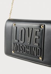 Μαύρη δερμάτινη τσάντα χειρός με ανάγλυφο κείμενο "LOVE MOSCHINO", χρυσές μεταλλικές λεπτομέρειες και λευκή ραφή. Ορθογώνιο σχήμα με λουρί αλυσίδα.