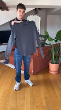 Jonge man die binnen staat en een lange mouwen grijze sweater vasthoudt in een woonkamer met houten vloer en planten.