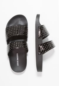 Des sandales noires à glisser avec deux larges sangles ornées de clous en argent, fabriquées dans un matériau brillant, avec une semelle texturée noire. Nom de la marque visible.