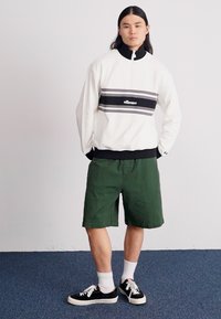 Witte zip-up sweatshirt met zwarte kraag, grijs en zwarte strepen, gecombineerd met groene shorts. Model draagt zwarte sneakers en witte sokken.