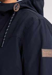 Granatowa kurtka z kapturem ze sznurkiem, brązowym zapięciem na guziki i teksturowaną naszywką z logo na rękawie. Gładki materiał z minimalnymi detalami.