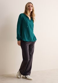 Felpa a maniche lunghe color teal con scollo a V, dotata di lacci, abbinata a pantaloni grigi scuri e sneakers bianche con dettagli neri.